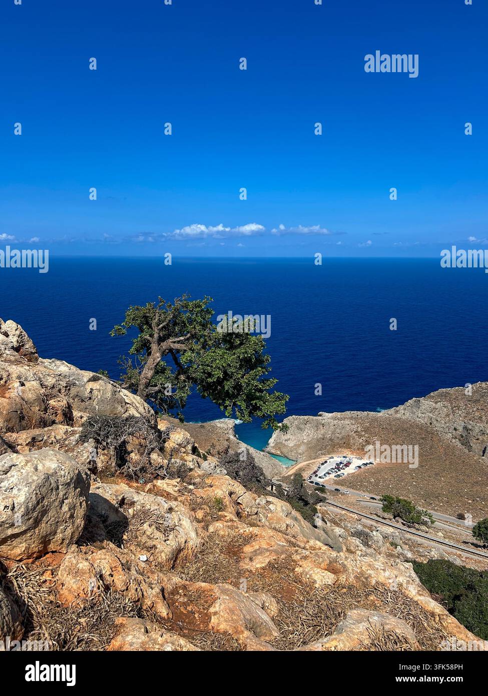Ein einsamer Baum klammert sich an eine felsige Klippe über dem tiefblauen Ägäischen Meer nahe Seitan Limania, mit einer sich windenden Küstenstraße unten, Kreta. - Smartphone-aufgenommenes Stockfoto