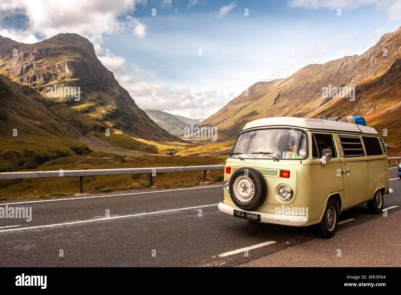 Ein klassischer Vintage-Campervan fährt eine malerische Straße durch Glen Coe in den schottischen Highlands entlang. Auf beiden Seiten des Tals erheben sich hohe Berggipfel unter einem hellen, teilweise bewölkten Himmel Stockfoto