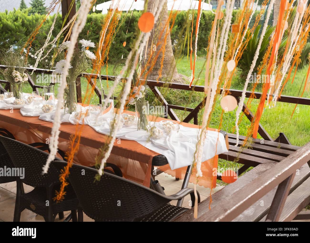 Tisch für eine Outdoor-Feier, dekoriert mit orangefarbenen und weißen hängenden Bändern, Glasgeschirr und Blumendetails in einer grünen Gartenumgebung. Stockfoto