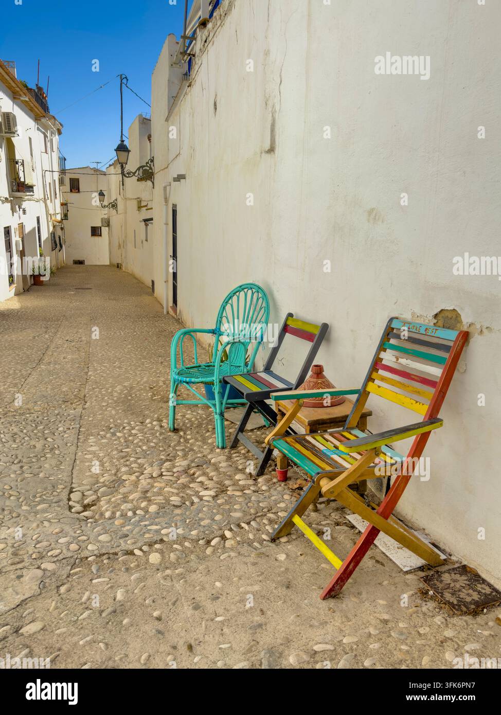 Bunte Stühle und ein kleiner Tisch säumen eine bezaubernde Kopfsteinpflasterstraße mit weißen Gebäuden unter einem klaren blauen Himmel in der historischen Altstadt von Altea. Stockfoto