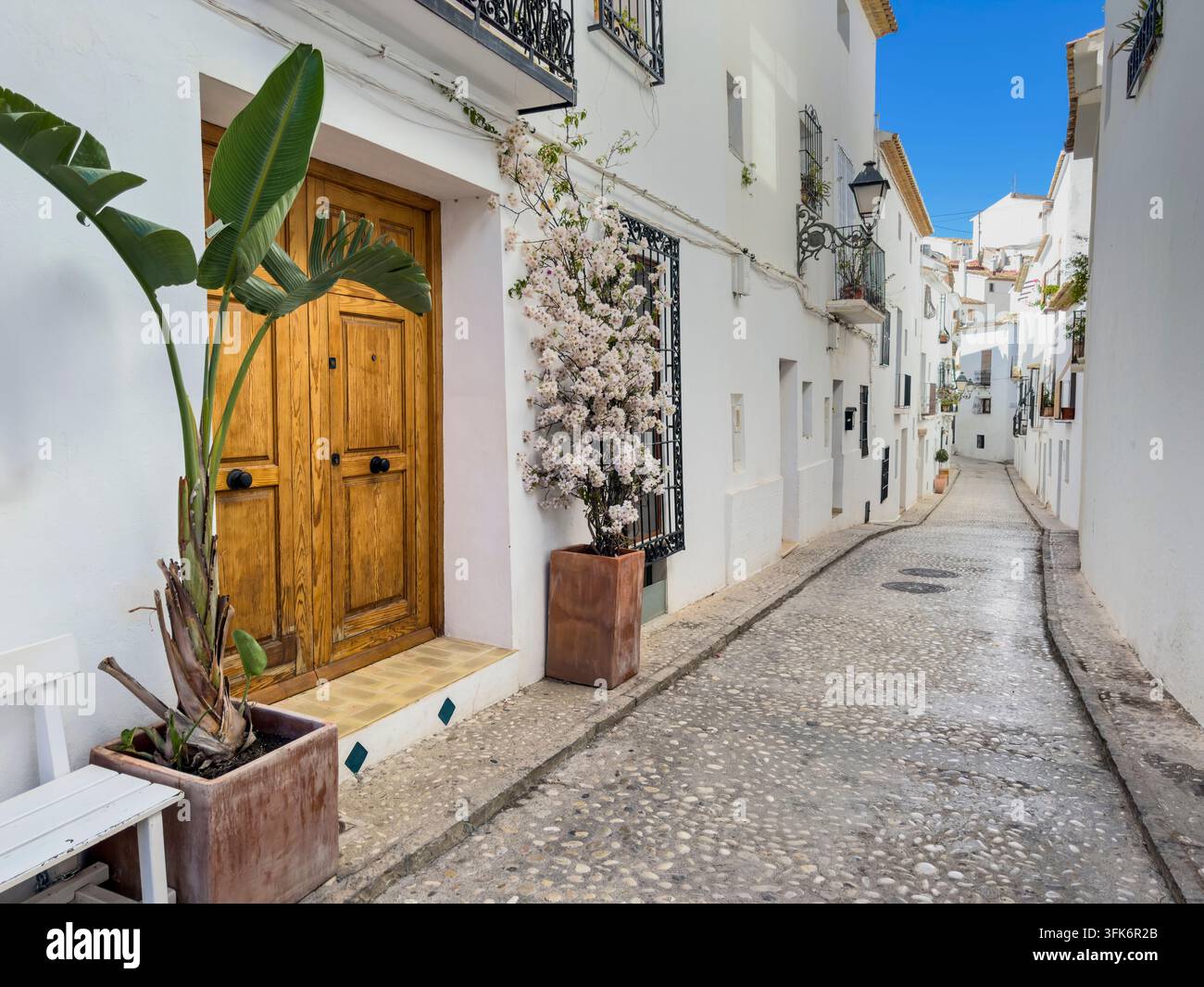 Altea-Straße mit weißen mediterranen Häusern entlang einer Kopfsteinpflastergasse, traditionelle Holztür flankiert von Topfpflanzen unter einem strahlend blauen Himmel. Stockfoto
