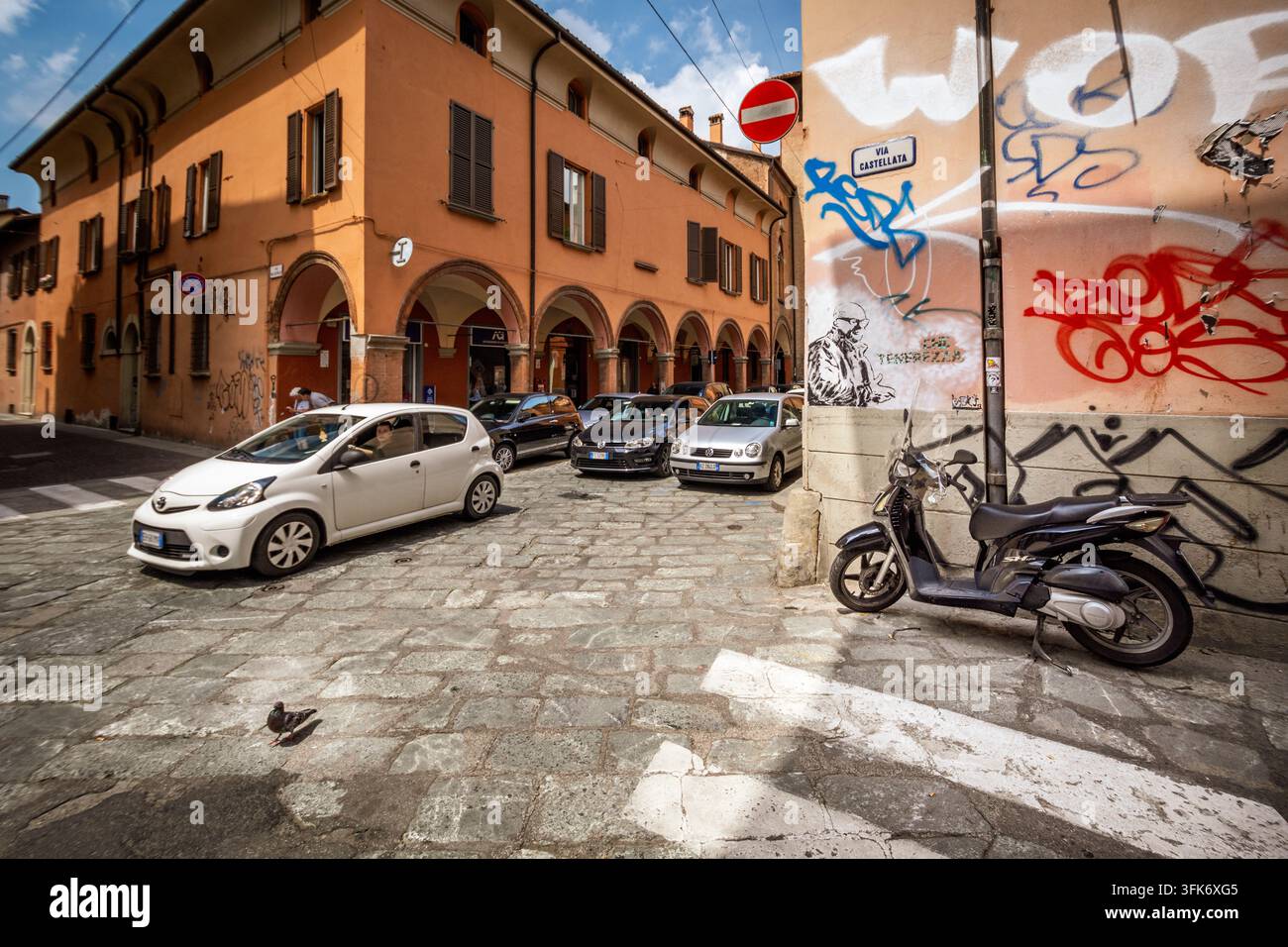 Bologna, Italien - 17. Mai 2018: Blick auf eine Kopfsteinpflasterstraße in Bologna mit historischen Säulenganggebäuden, geparkten Autos, einem Roller und Graffiti auf der C Stockfoto