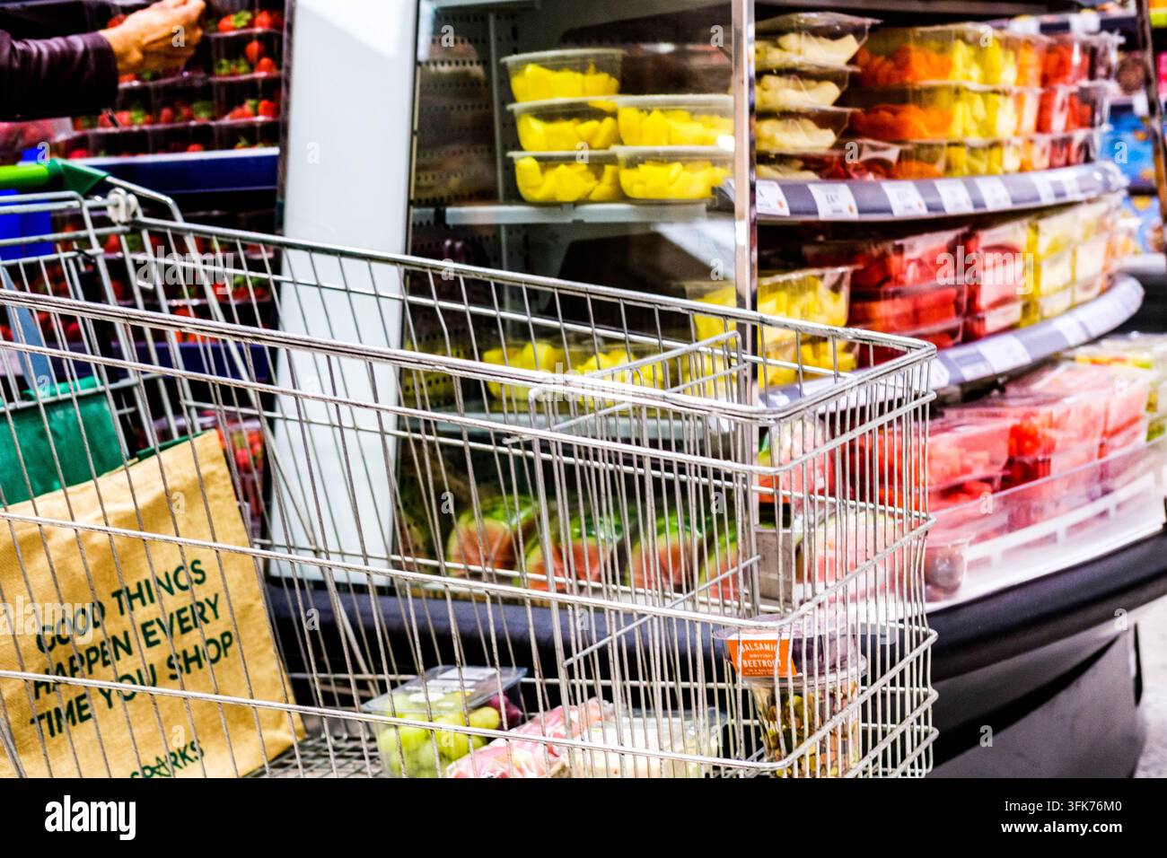 London, UK, 23. September 2026, Einkaufswagen mit frischen Trauben und vorgepacktem Salat vor farbenfrohem mehrstufigem Display von tropischen Obsttöpfen Stockfoto