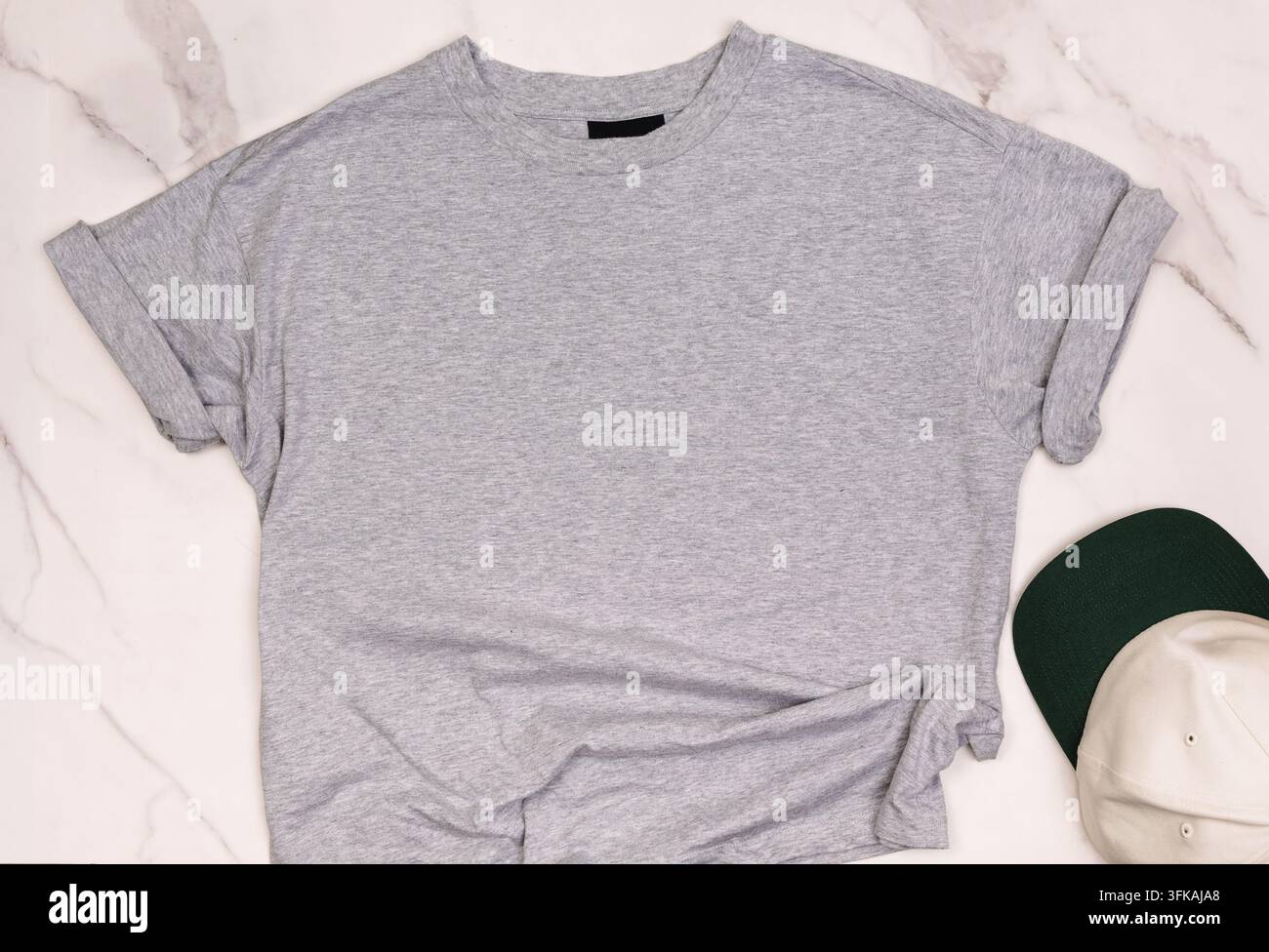 Leere graue Oversized-T-Shirt mit aufgerollten Ärmeln und einem leeren Halsetikett auf weißem Marmor neben einer Baseballkappe, Ansichtsmodell von oben Stockfoto