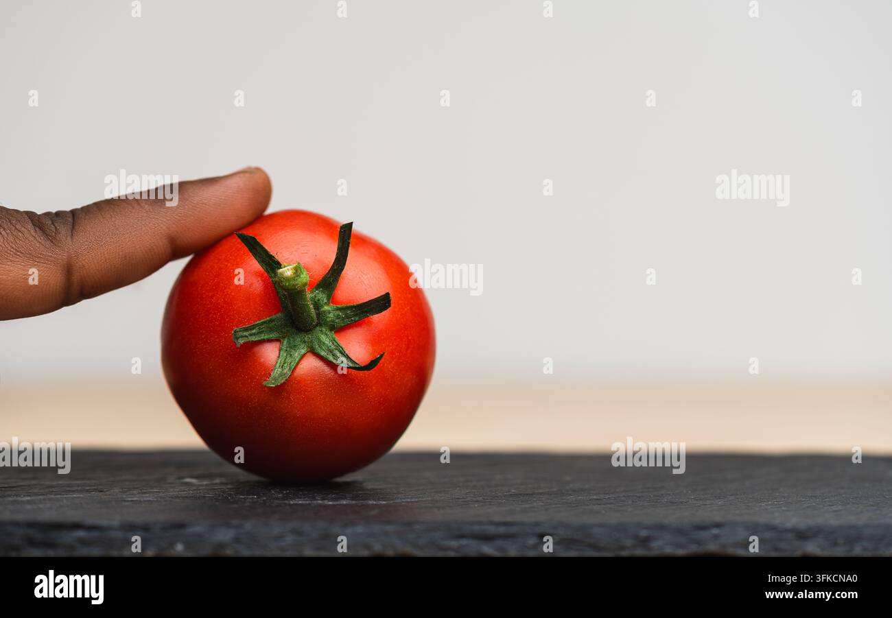 Finger tip berührt eine frische reife rote Tomate auf einer dunklen Schieferplatte vor einem hellen Hintergrund, minimales Lebensmittelkonzept mit Kopierraum Stockfoto