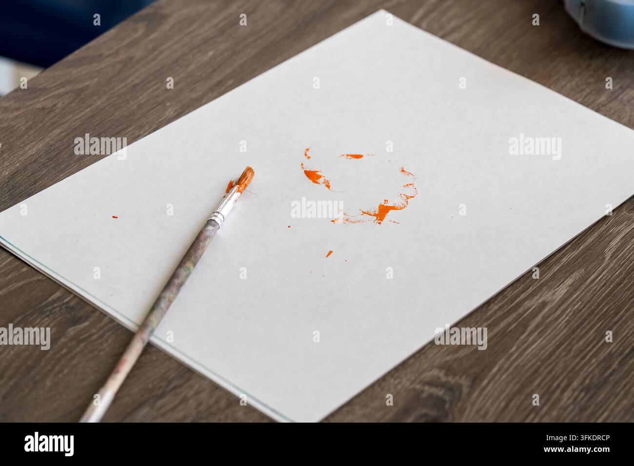 Weißes Papier mit orangefarbenen Farbflecken und einem gebrauchten Pinsel auf einem Holztisch. Kreativer Kunstprozess, Malerei oder DIY-Bastelkonzept. Stockfoto