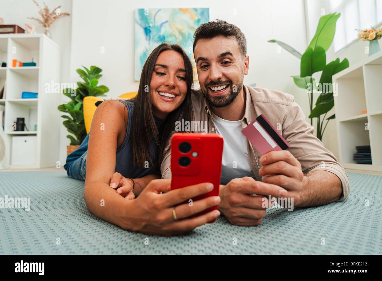 Ein fröhliches Paar liegt auf einem weichen Teppich, beschäftigt mit einem roten Smartphone und einer Kreditkarte, und genießt einen gemeinsamen digitalen Moment und spielerische Entdeckung zusammen. Stockfoto