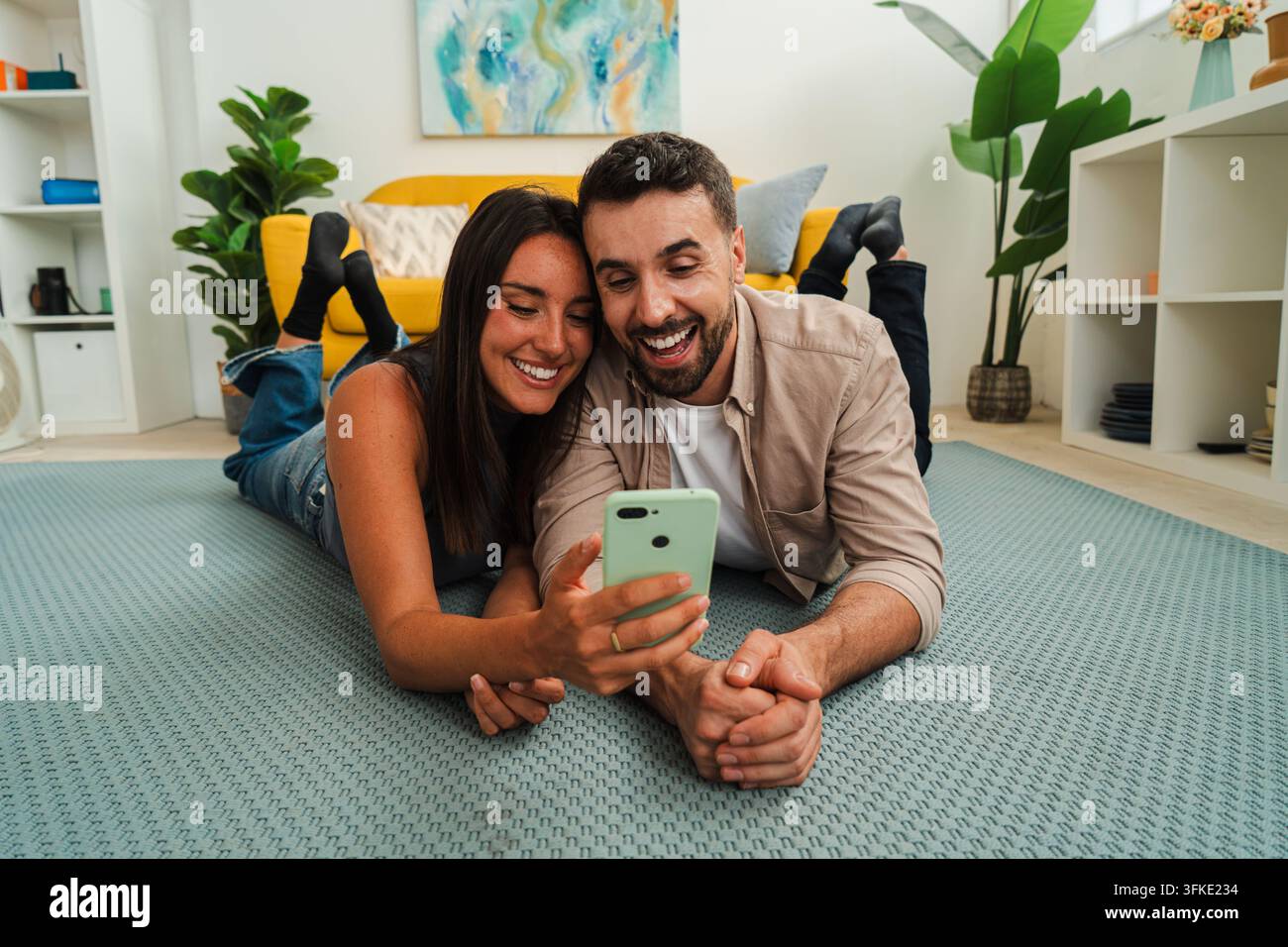 Zwei Personen teilen einen ungezwungenen Moment, während sie einen Smartphone-Bildschirm betrachten und zusammen auf einem weichen Teppich in einem hellen Zuhause liegen. Warmes Licht und entspannte Stimmung Stockfoto