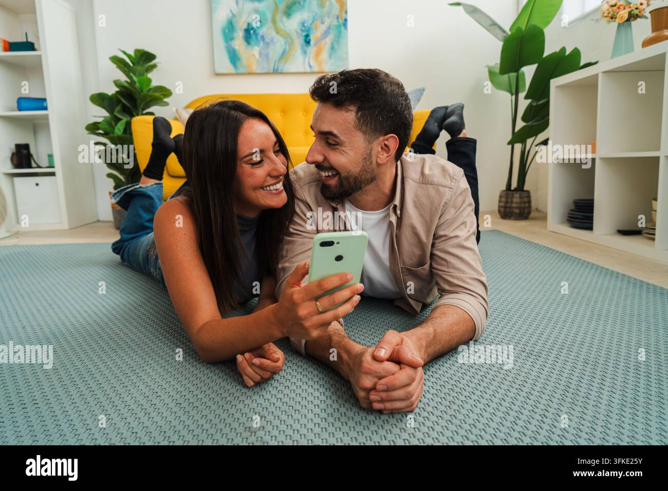 Zwei Personen genießen einen entspannten Moment auf einem weichen Teppich und erkunden Inhalte auf einem gemeinsamen Handy. Ihr warmes Lächeln und ihre enge Haltung vermitteln Verbundenheit und Neugier Stockfoto