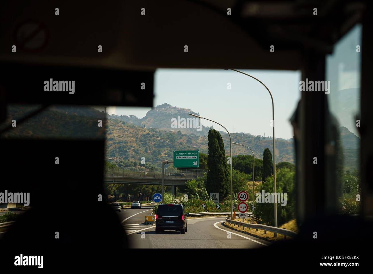 Giardini Naxos, Italien - 26. Juni 2026: Blick durch ein Busfenster, der eine malerische Autofahrt mit Berglandschaft und Verkehrsschildern auf einer sonnigen Autobahn unter blauem Himmel zeigt, Reiseabenteuer und Roadtrip-Atmosphäre Stockfoto