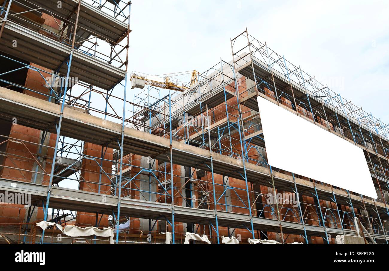 Leere Informations-tafel an der Fassade eines neuen großen Gebäudes im Bau zwischen den Gerüsten. Kran und bewölkter Himmel darüber. Hintergrund für Text Stockfoto