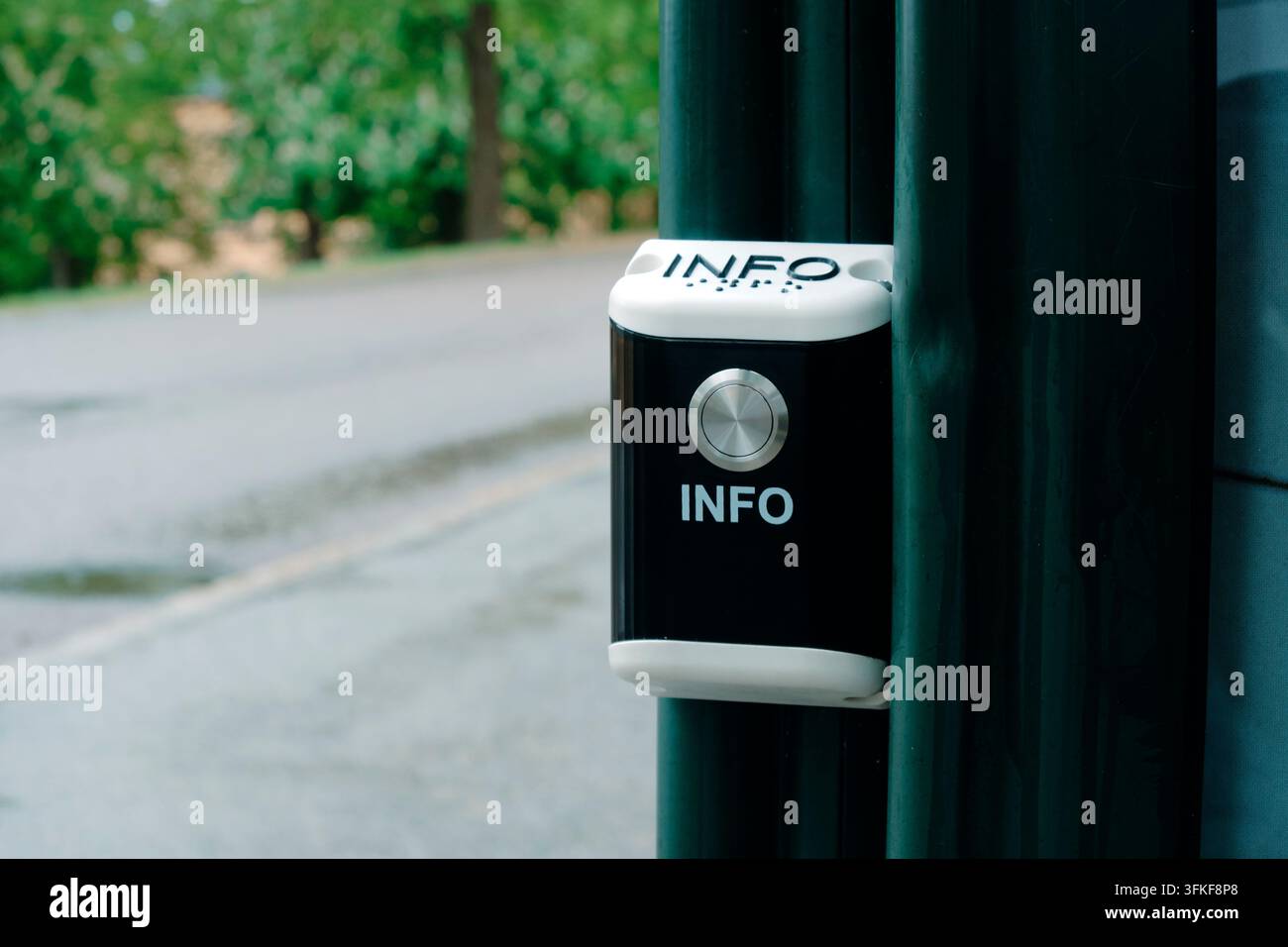 neben einem gepflasterten Weg ist ein öffentlicher Informationsknopf mit Braille-Schrift und einem großen silbernen Druckknopf an einem dunkelgrünen Metallpfosten befestigt, wi Stockfoto