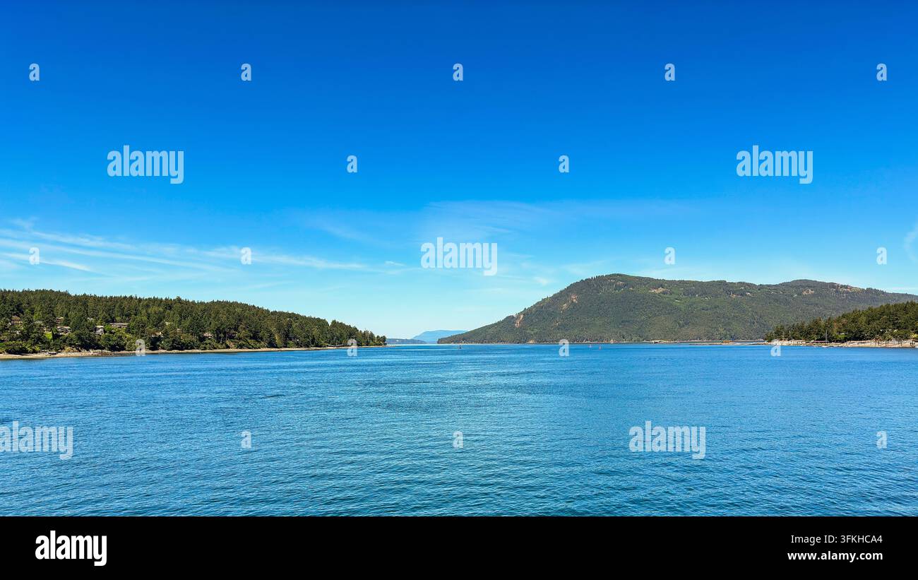 Ein markanter Hügel auf der rechten Seite blickt auf ruhige Küstengewässer und einen klaren blauen Himmel. Stockfoto