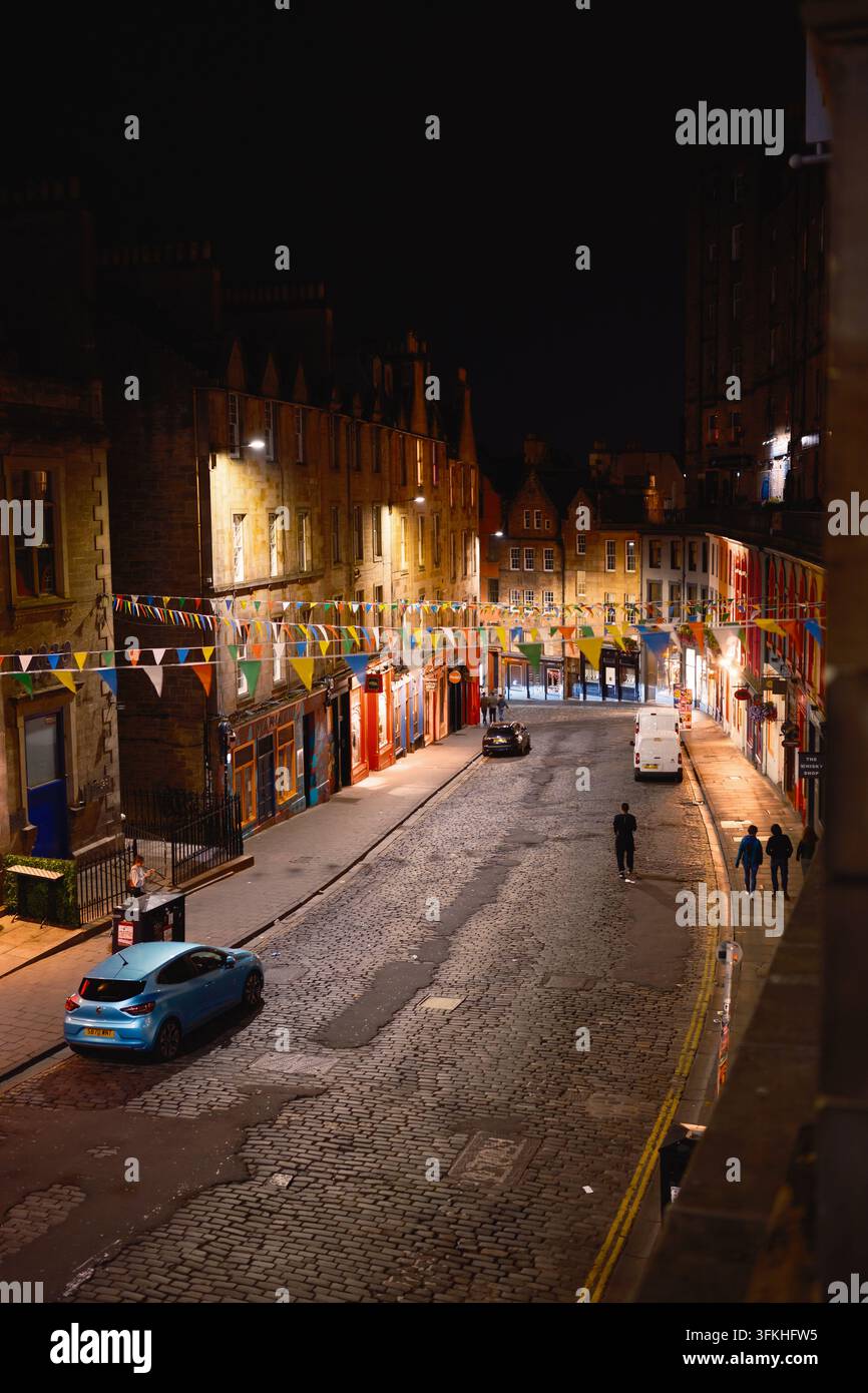 Edinburgh, Schottland – 3. August 2026: Nachtansicht einer historischen Kopfsteinpflasterstraße mit bunten Fahnen, warmem Licht, geparkten Autos und Fußgängern. Stockfoto