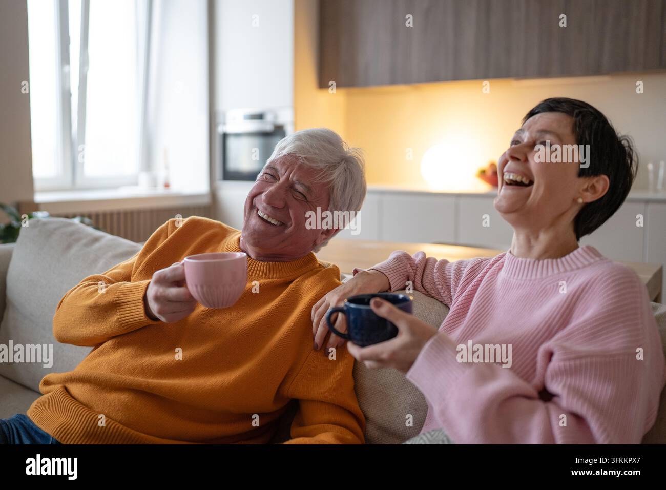 Fröhliches Seniorenpaar lacht zusammen mit Teetassen, entspannt auf dem Sofa zu Hause im Ruhestand Stockfoto