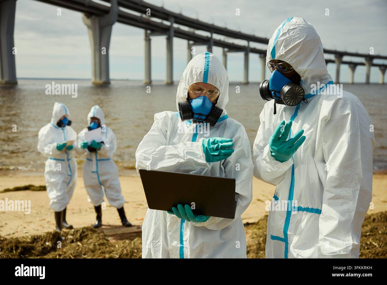 Verschiedene Ökologen in Schutzanzügen, die einen Laptop an einer verschmutzten Küste verwenden Stockfoto
