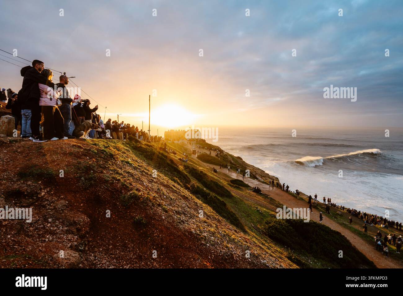 Nazare, Portugal - 6. Dezember 2025: Menschen stehen auf Klippen und beobachten den Ozean und die großen Wellen bei Sonnenuntergang in Nazare, Portugal Stockfoto