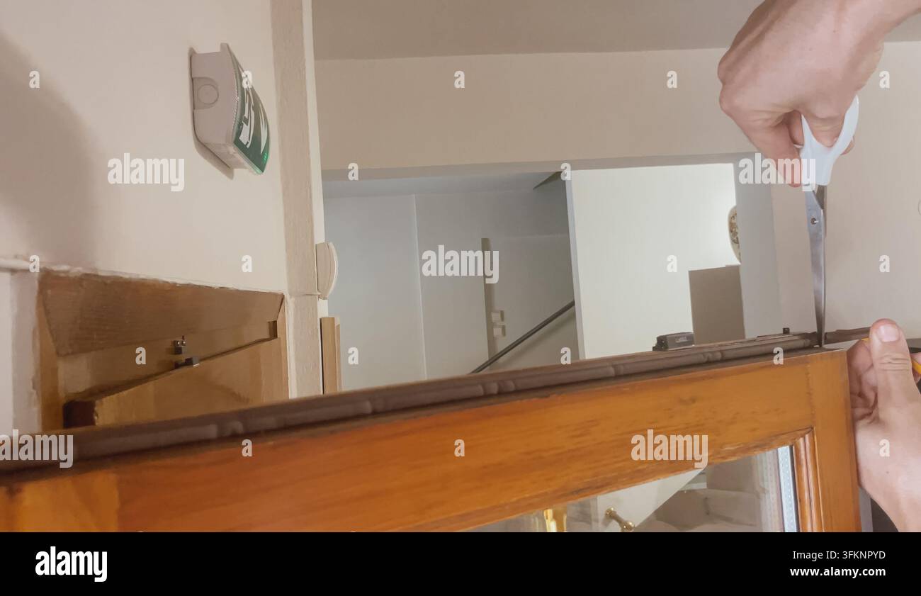 Horizontale Nahaufnahme von Männerhänden, die selbstklebendes Gummidichtband auf einen Holzfensterrahmen für Heimdämmung und Schalldämmung anbringen Stockfoto