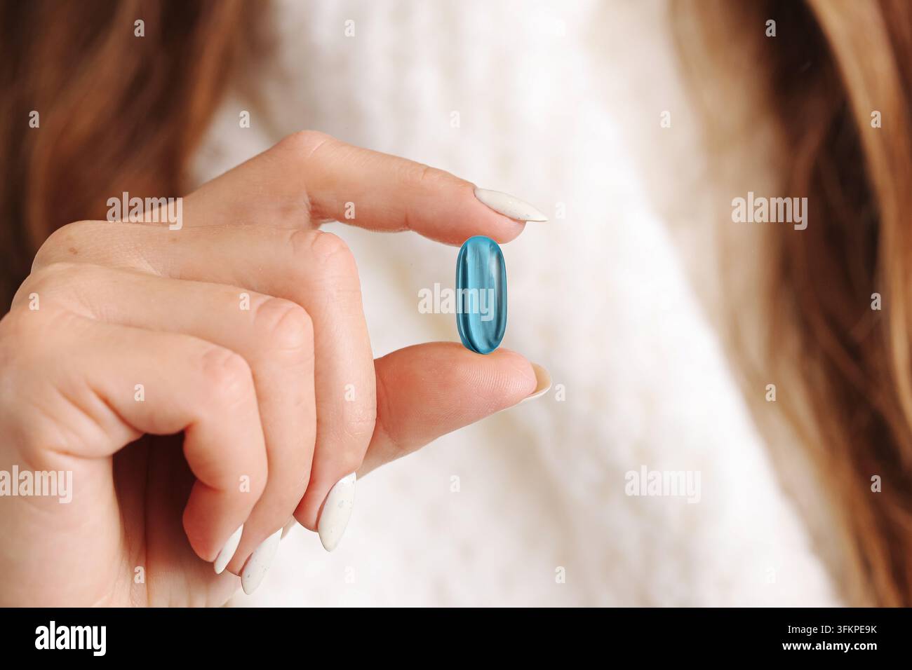 Blaue Nahrungsergänzungsmittel-Kapsel, die in einer Lifestyle-Szene nahe dem Gesicht einer Frau gehalten wird. Wellness-Hintergrund für tägliche Vitamnernährung, Selbstpflege und gesunde Gewohnheiten. Stockfoto
