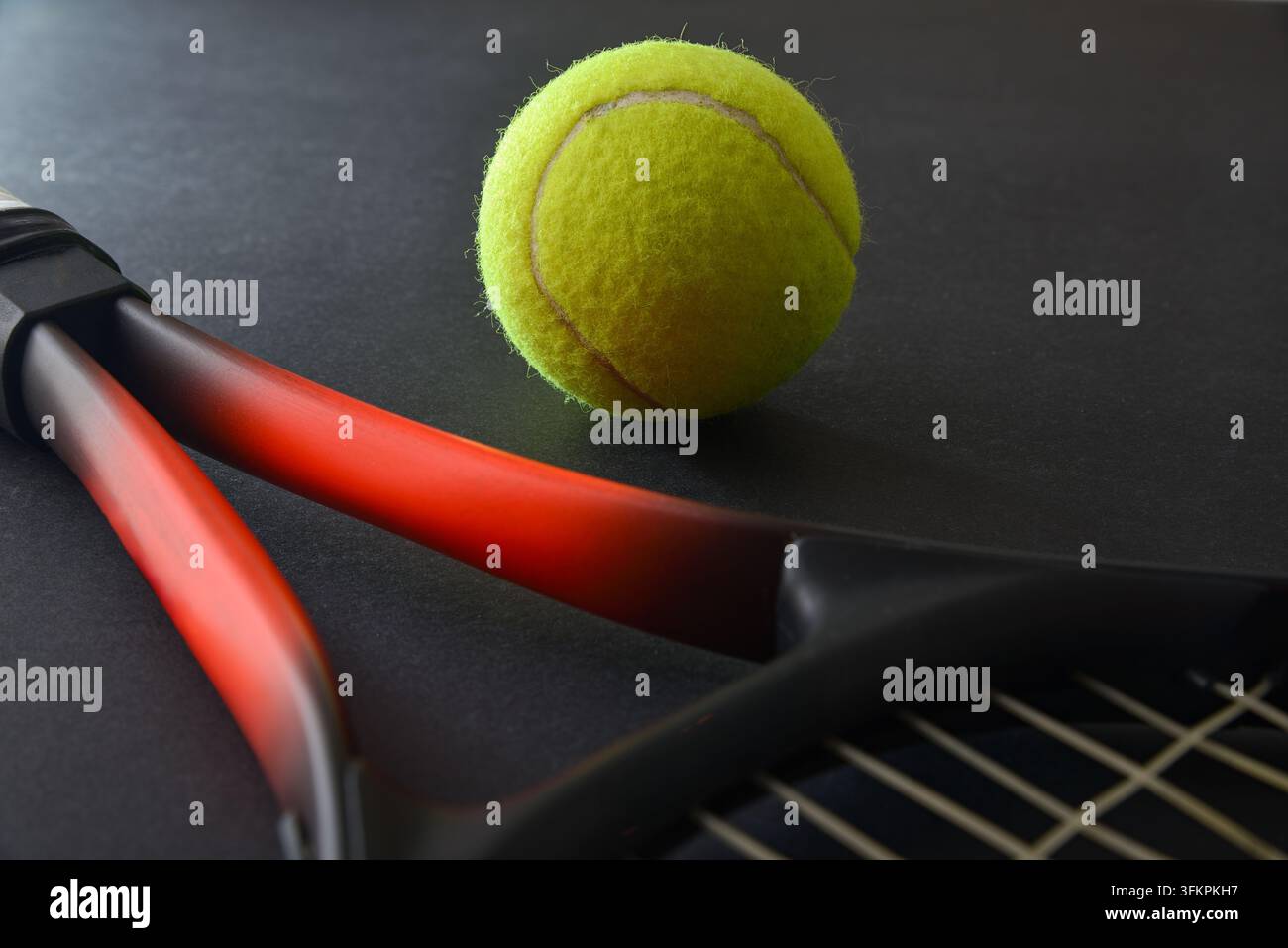 Ein hellgelber Tennisball ruht neben einem rot-schwarzen Schläger auf einer dunklen Oberfläche und schafft ein dramatisches Sportstillleben mit starkem Kontrast. Stockfoto