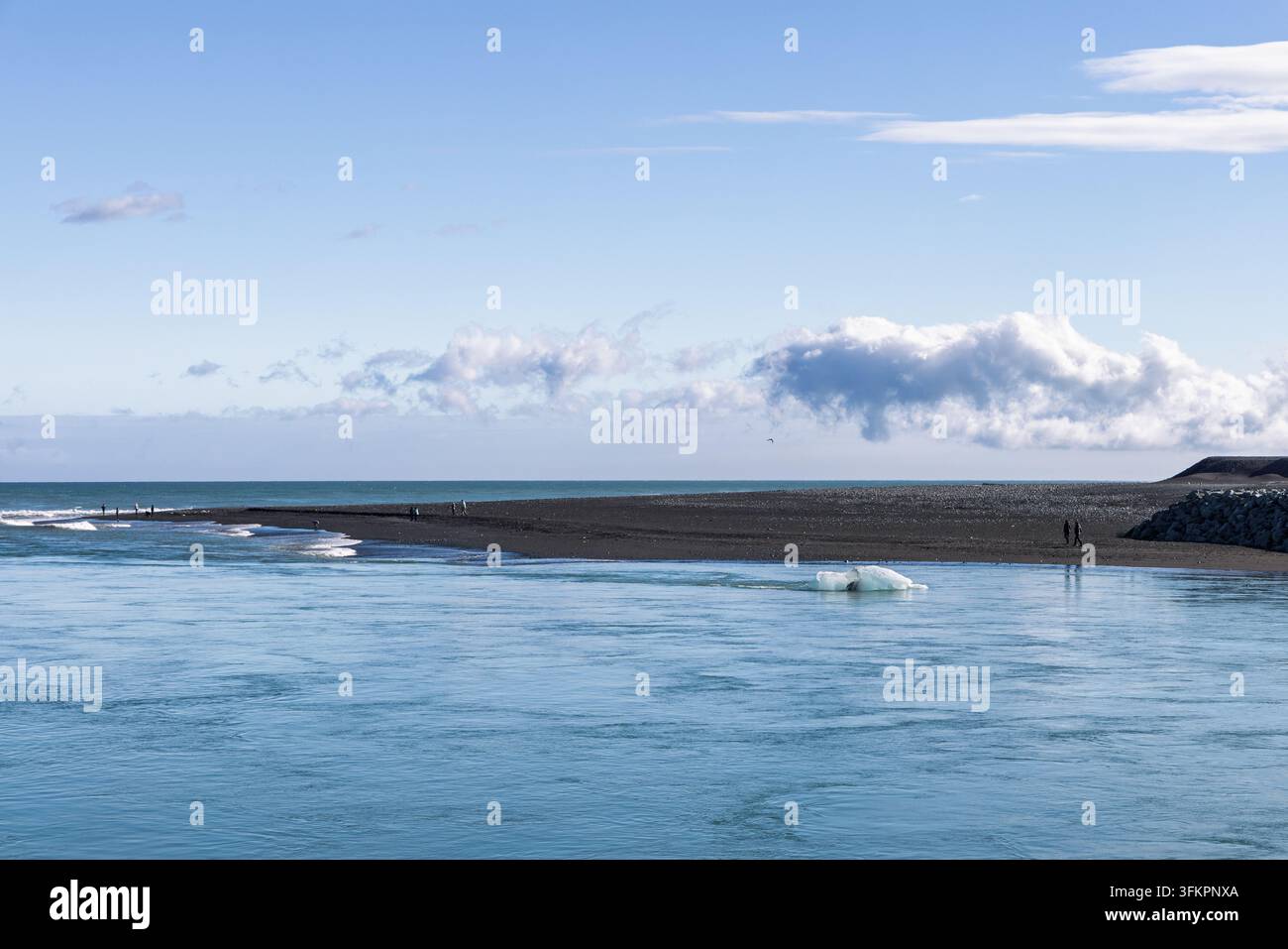 Schwebendes Eis in der Nähe einer schwarzen Sandzunge am Flussauslauf des Jökulsárlon, Diamond Beach, Island, mit entfernten Besuchern und weitem blauem Himmel. Stockfoto