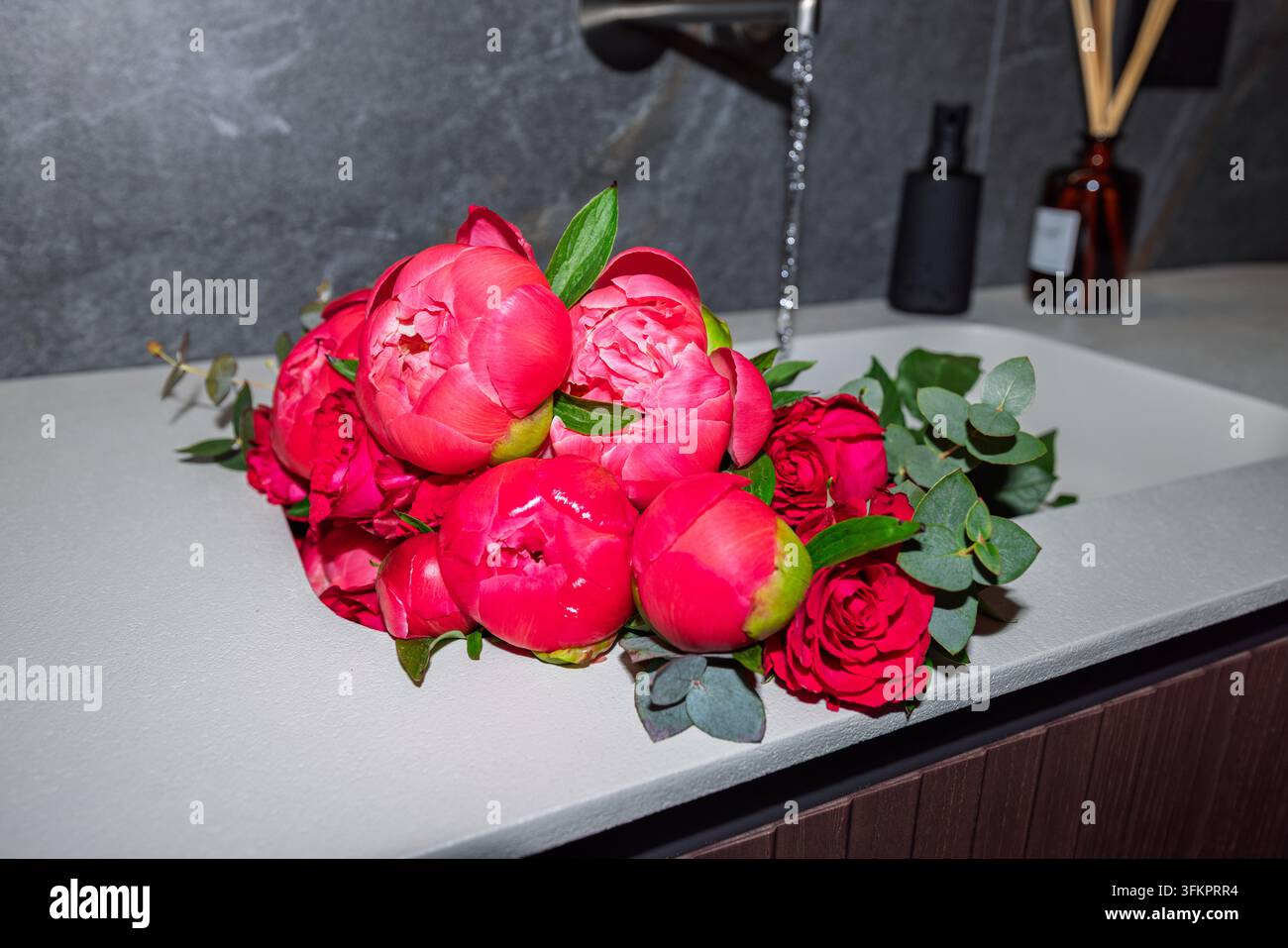 Rosa Pfingstrosen und rote Rosen füllen ein weißes Waschbecken, während Wasser aus einem schwarzen Wasserhahn dahinter in einem zeitgenössisch italienisch gestalteten Badezimmer fließt. Stockfoto