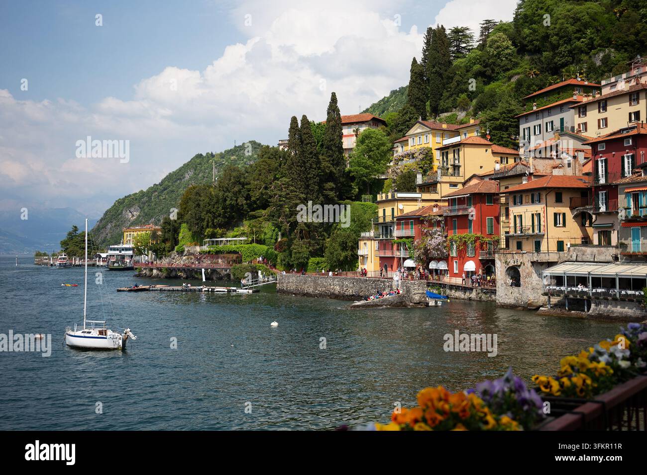 Die roten und gelben Uferhäuser von Varenna rahmen ein weißes Segelboot auf blauem Wasser ein, mit farbenfrohen blühenden Stiefmütterchen auf der Promenade im Vordergrund Stockfoto