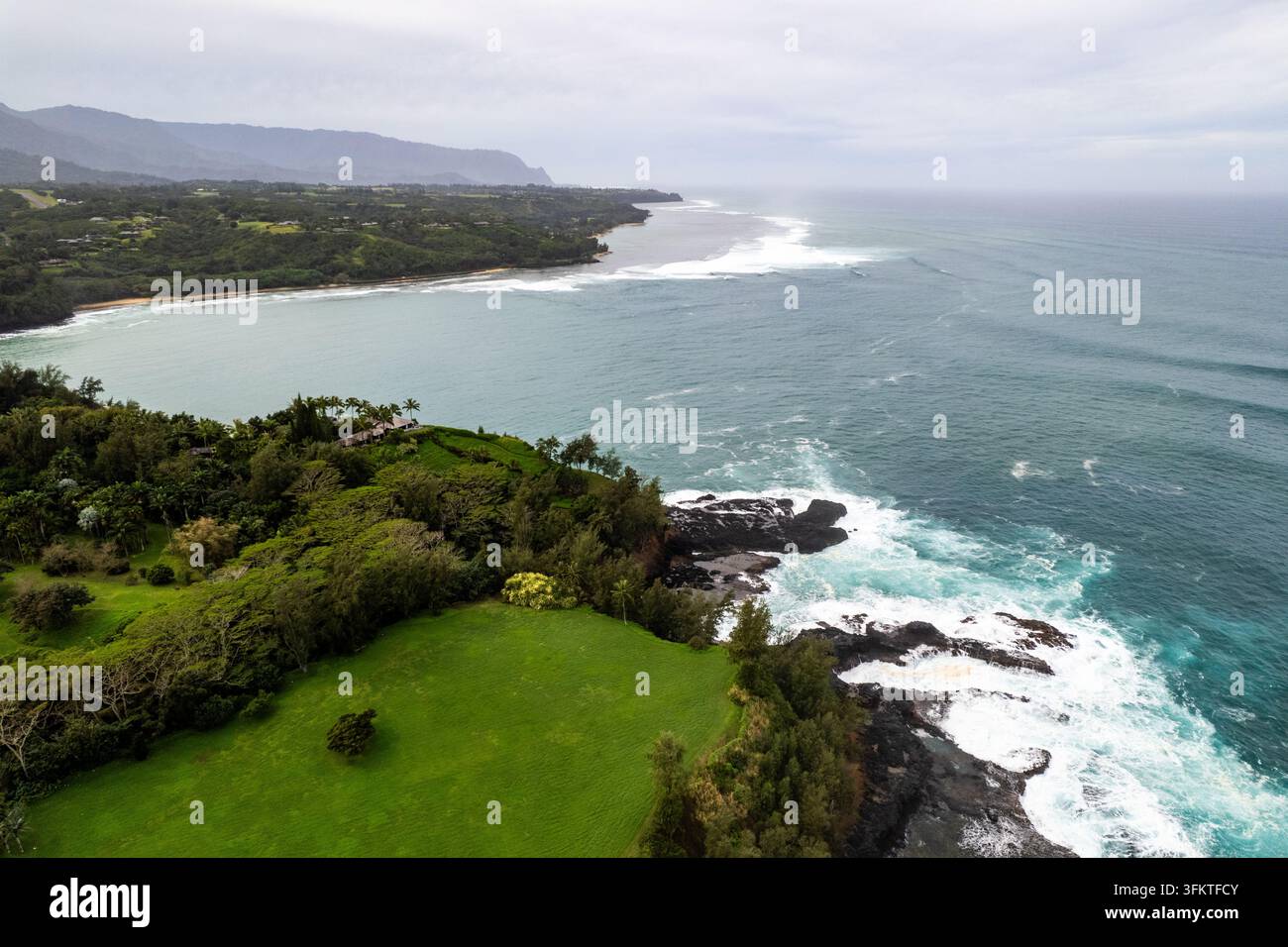 Oberblick auf die Landschaft von Kauai mit einem üppigen grünen Fairway, das zu einer dunklen vulkanischen Küste führt, wo die Pazifikwellen hereinrollen, mit felsigem schwarzem Lava und Cloß Stockfoto