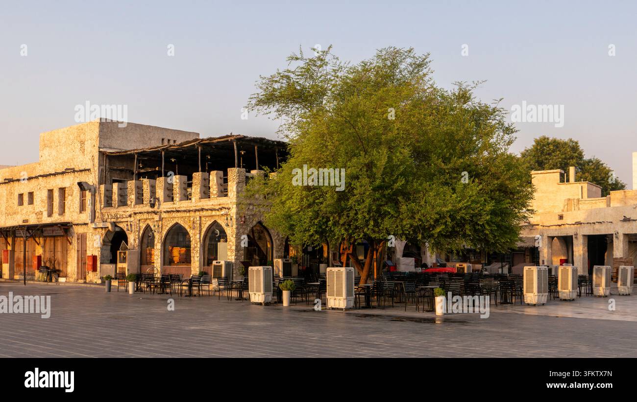 Außenbestuhlung eines Restaurants, beschattet von einem belaubten grünen Baum neben traditioneller Steinarchitektur im Souq Waqif in Doha, Katar. Stockfoto