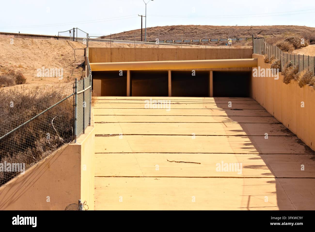Ein trockener Betonüberlaufkanal verläuft unter einer Straße zwischen eingezäunten, sonnenverbrannten Hängen unter der hellen Sommersonne in Riverside, Kalifornien. Stockfoto