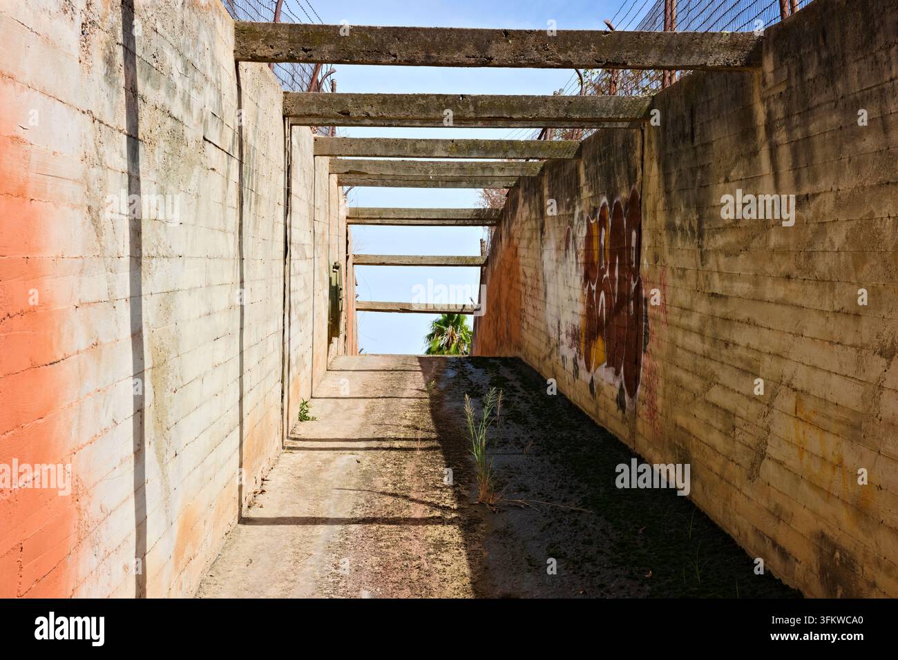 Verwitterte Betonwände und sich wiederholende Überkopfträger bilden einen schmalen trockenen Kanal mit tiefen Schatten und spärlichem Unkraut in Riverside, Kalifornien. Stockfoto