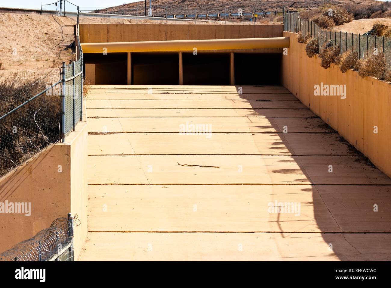Ein trockener Betonüberlaufkanal verläuft unter einer Straße zwischen eingezäunten, sonnenverbrannten Hängen unter der hellen Sommersonne in Riverside, Kalifornien. Stockfoto