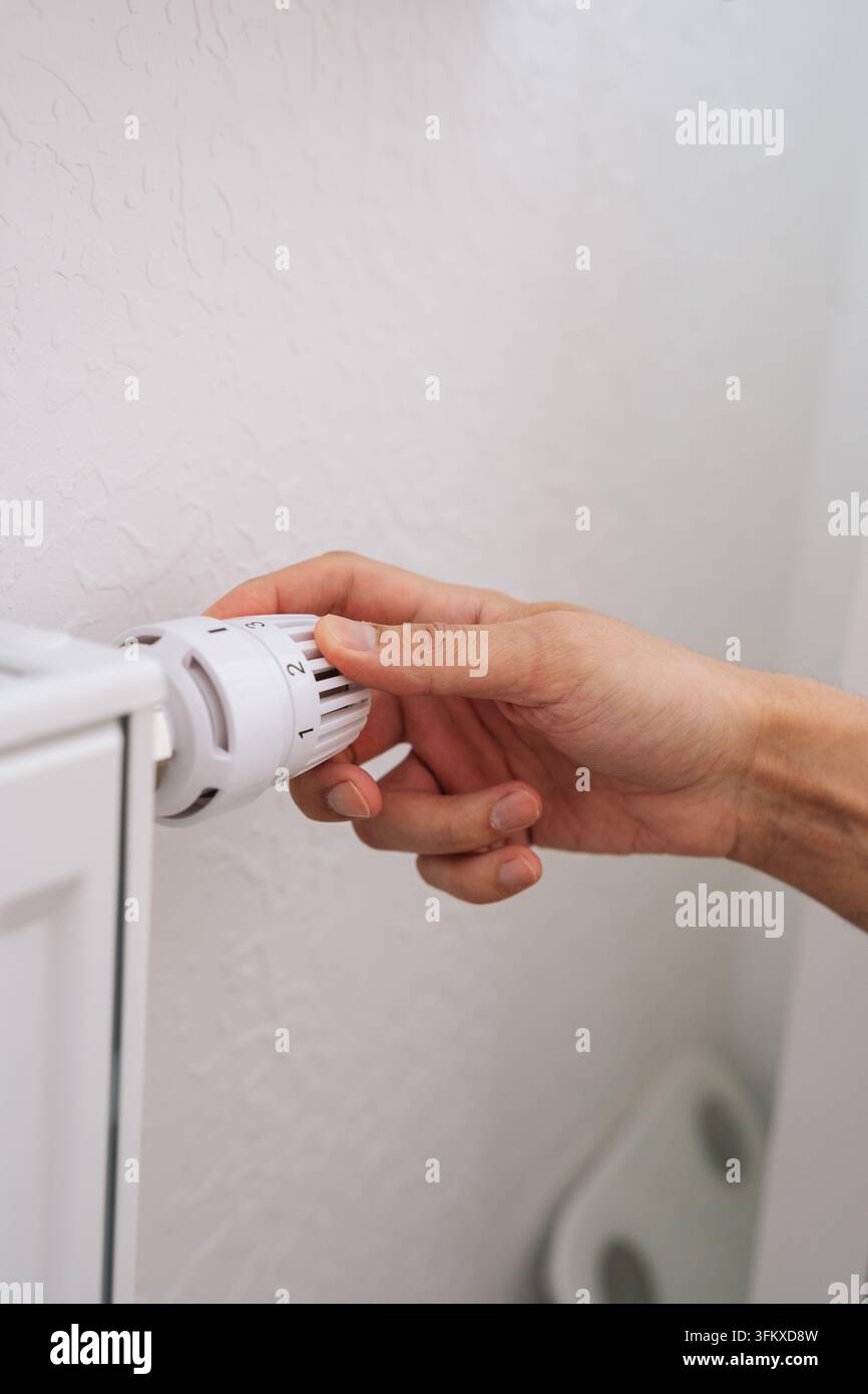 Nahaufnahme einer Hand, die den weißen Thermostatknopf des Heizkörpers einstellt. Konzept für Energieeinsparung, Kontrolle der Zimmertemperatur und Reduzierung der Heizkosten. Minima Stockfoto