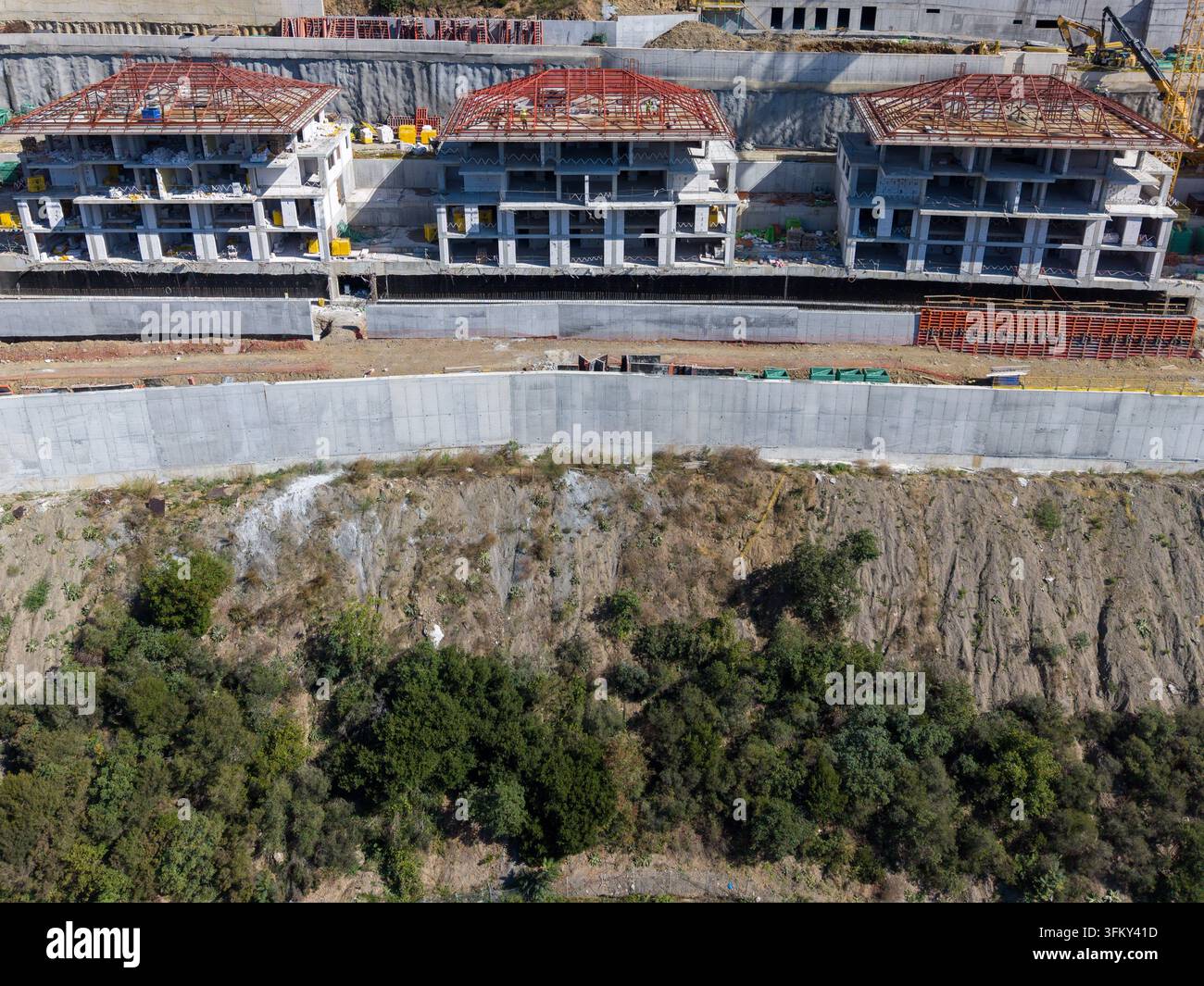 Luftaufnahme einer Baustelle mit drei modernen Wohngebäuden mit roten Dachrahmen, einer großen Beton-Stützmauer und einem Hügel Stockfoto