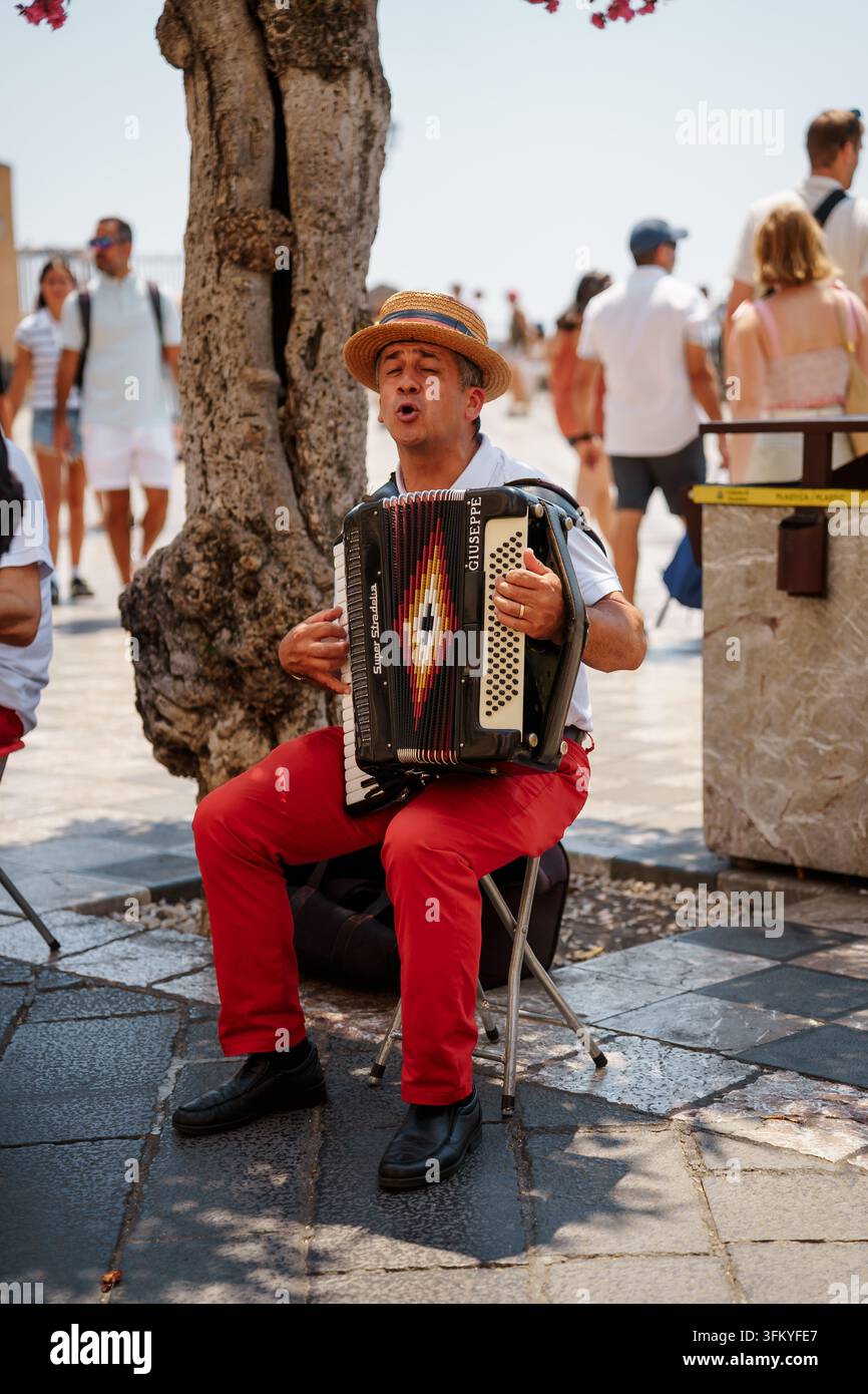 Taormina, Italien - 26. Juni 2026: Straßenmusiker spielt eine Akkordeon im Freien unter einem Baum, trägt einen Strohhut und eine rote Hose. Lebendige Szene von Kulturmusik und Tourismus, sonnige Atmosphäre Stockfoto
