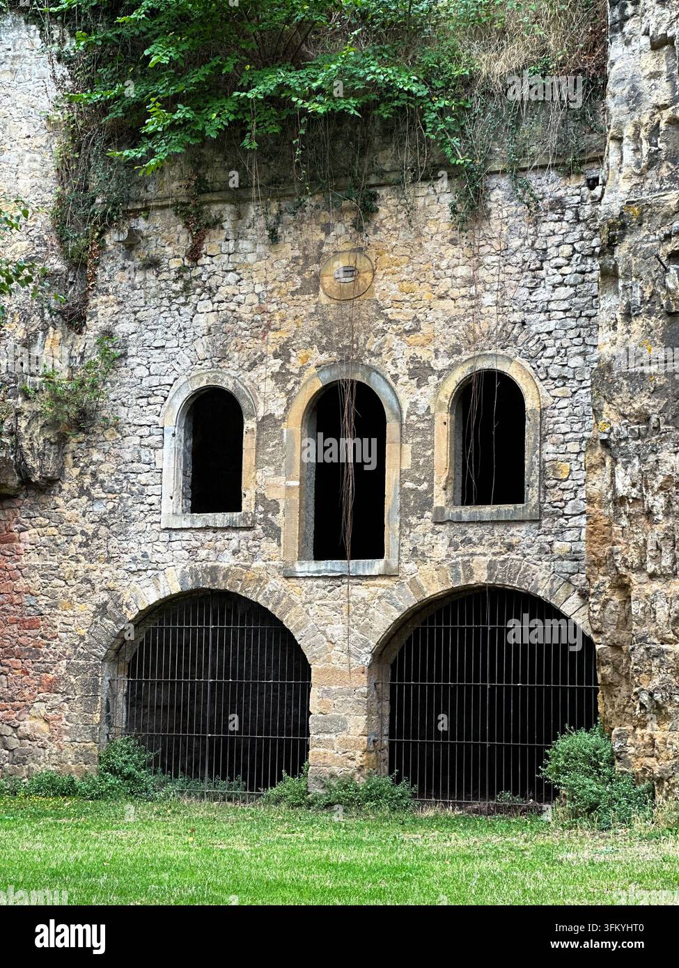 Eine alte Steinfortificationsmauer zeigt drei gewölbte obere Fenster und zwei große vergitterte untere Tore unter einer überwucherten Felswand. - Smartphone-aufgenommenes Stockfoto