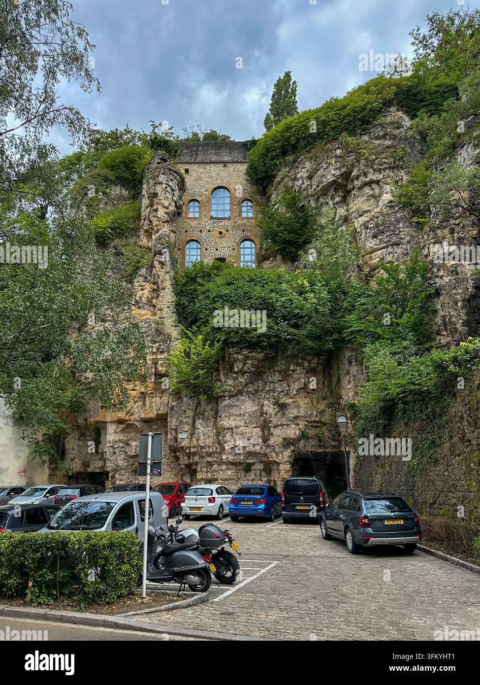 Ein altes Steingebäude ist in eine Felswand eingebaut, umgeben von grünen Blattwerk, mit Autos, die entlang des Kopfsteinpflasters unten geparkt sind. - Smartphone-aufgenommenes Stockfoto