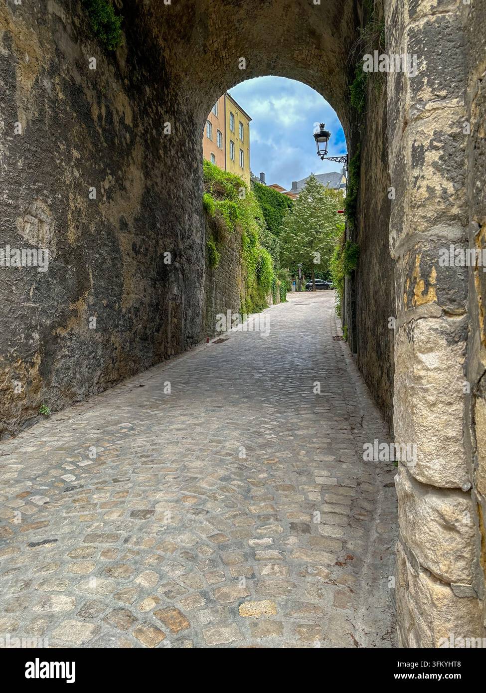 Alter steinerner Torbogen, der eine malerische historische Straße einrahmt. Alter steinerner Torbogen, der eine gepflasterte Straße einrahmt, die zu historischen Gebäuden und üppigem Grün führt Stockfoto