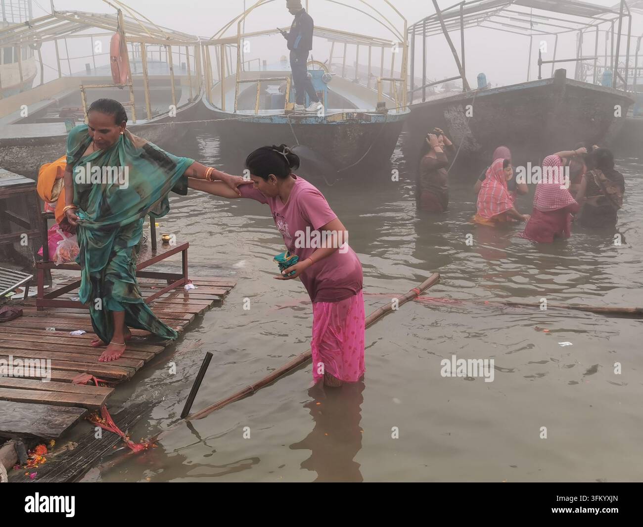VARANASI, INDIEN - 23. JANUAR 2026: Frauen helfen sich gegenseitig beim Baden im Ganges zwischen festgemachten Booten an einem nebligen Morgen. Stockfoto