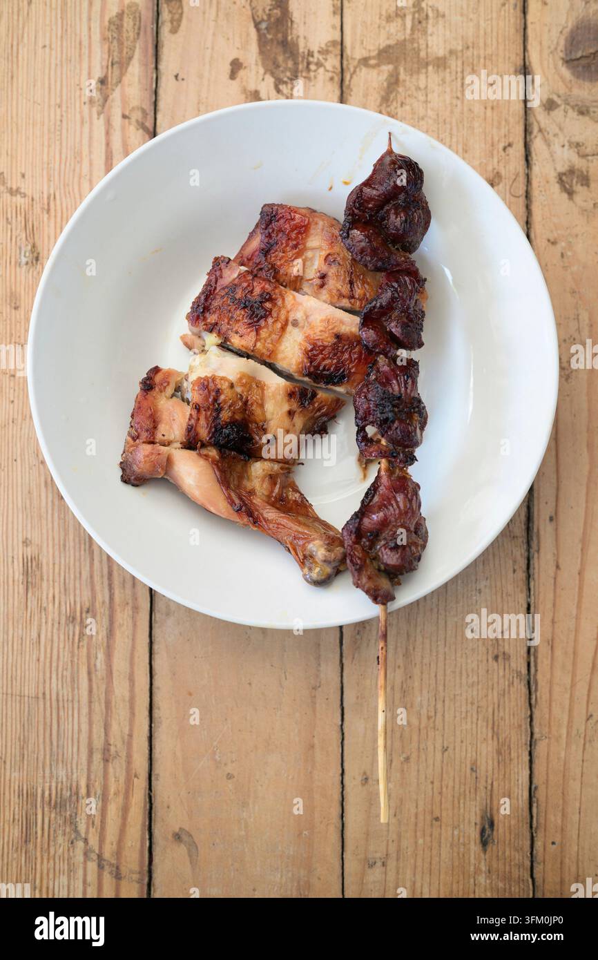 Eine Aufsicht auf das thailändische Street-Food-Grillhuhn und einen Leberspieß auf einem weißen Keramikteller. Das Tablett steht auf einem rustikalen hellen Holz Stockfoto