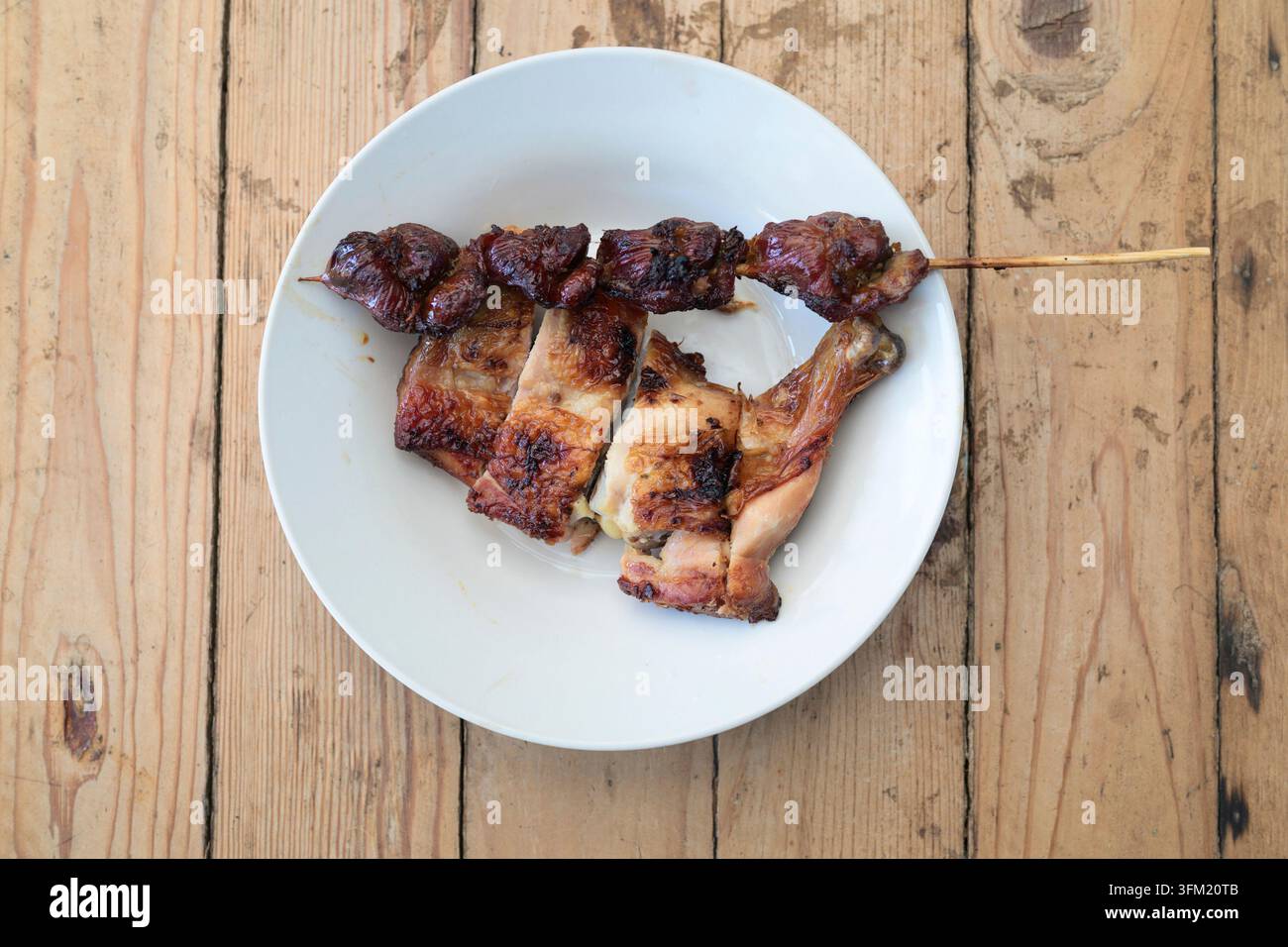 Eine Aufsicht auf das thailändische Street-Food-Grillhuhn und einen Leberspieß auf einem weißen Keramikteller. Das Tablett steht auf einem rustikalen hellen Holz Stockfoto