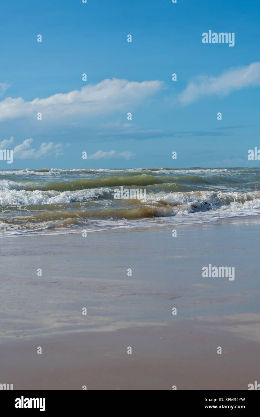 Dieses malerische Foto zeigt eine lebendige Meereslandschaft mit rollenden Wellen, die unter einem strahlend blauen Himmel, der mit flauschigen weißen Wolken übersät ist, auf einen sandigen Strand krachen Stockfoto
