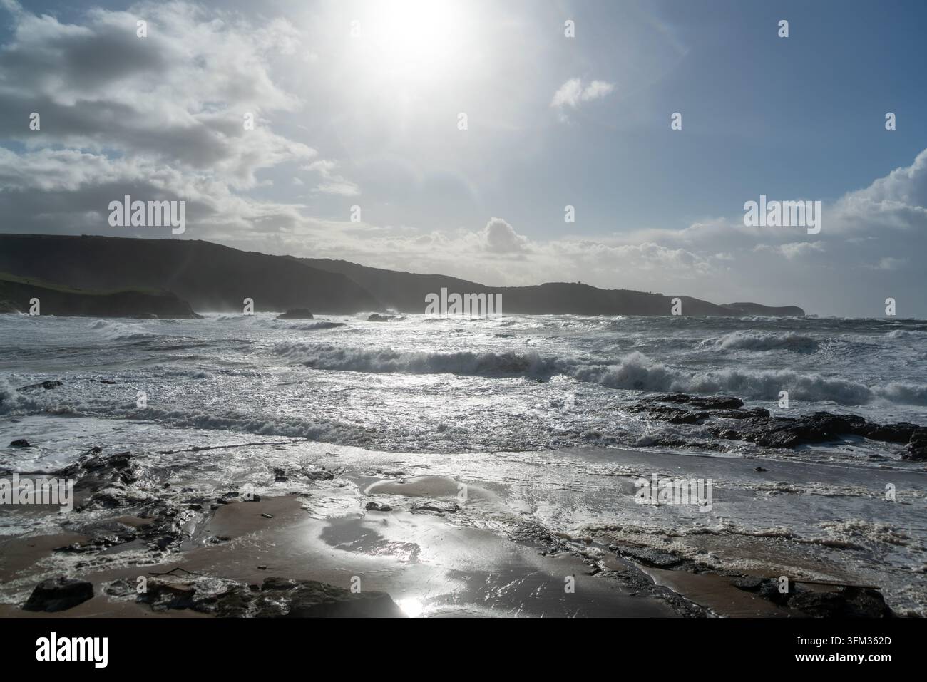 Dieses weitläufige Foto fängt eine dramatische Küstenszene ein und zeigt mächtige Wellen, die unter einem teilweise bewölkten Himmel gegen einen Sandstrand schlagen. Stockfoto