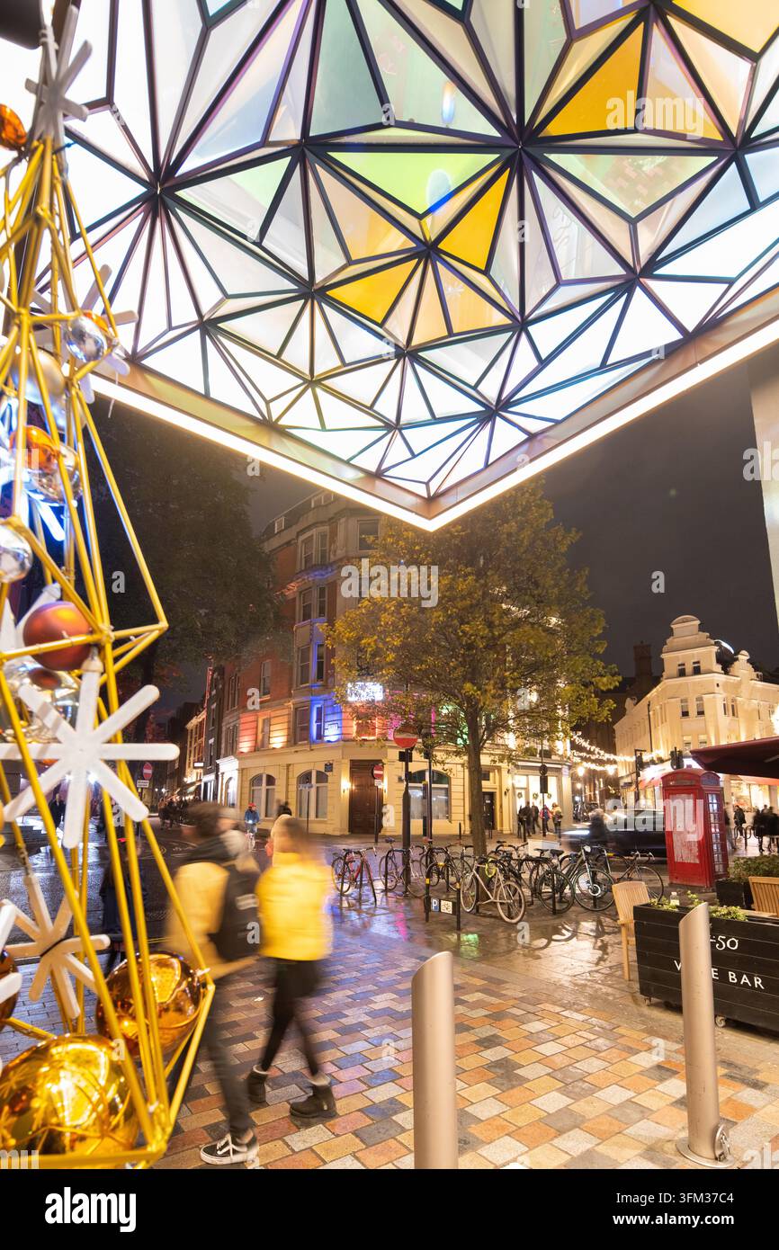 Menschen laufen unter dem farbenfrohen geometrischen Glasdach der Nova Victoria in London und genießen die abendlichen Weihnachtslichter. Stockfoto