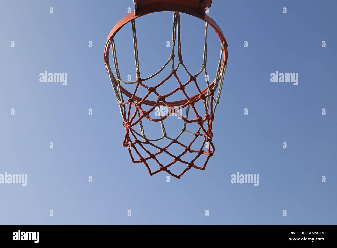 Von einem niedrigen Winkel aus aufgenommen, heben sich der orangefarbene Basketballkorb und das rot-weiße Netz vor dem klaren blauen Himmel ab. Das Sonnenlicht wirft einen warmen Schimmer und zeigt die simp Stockfoto