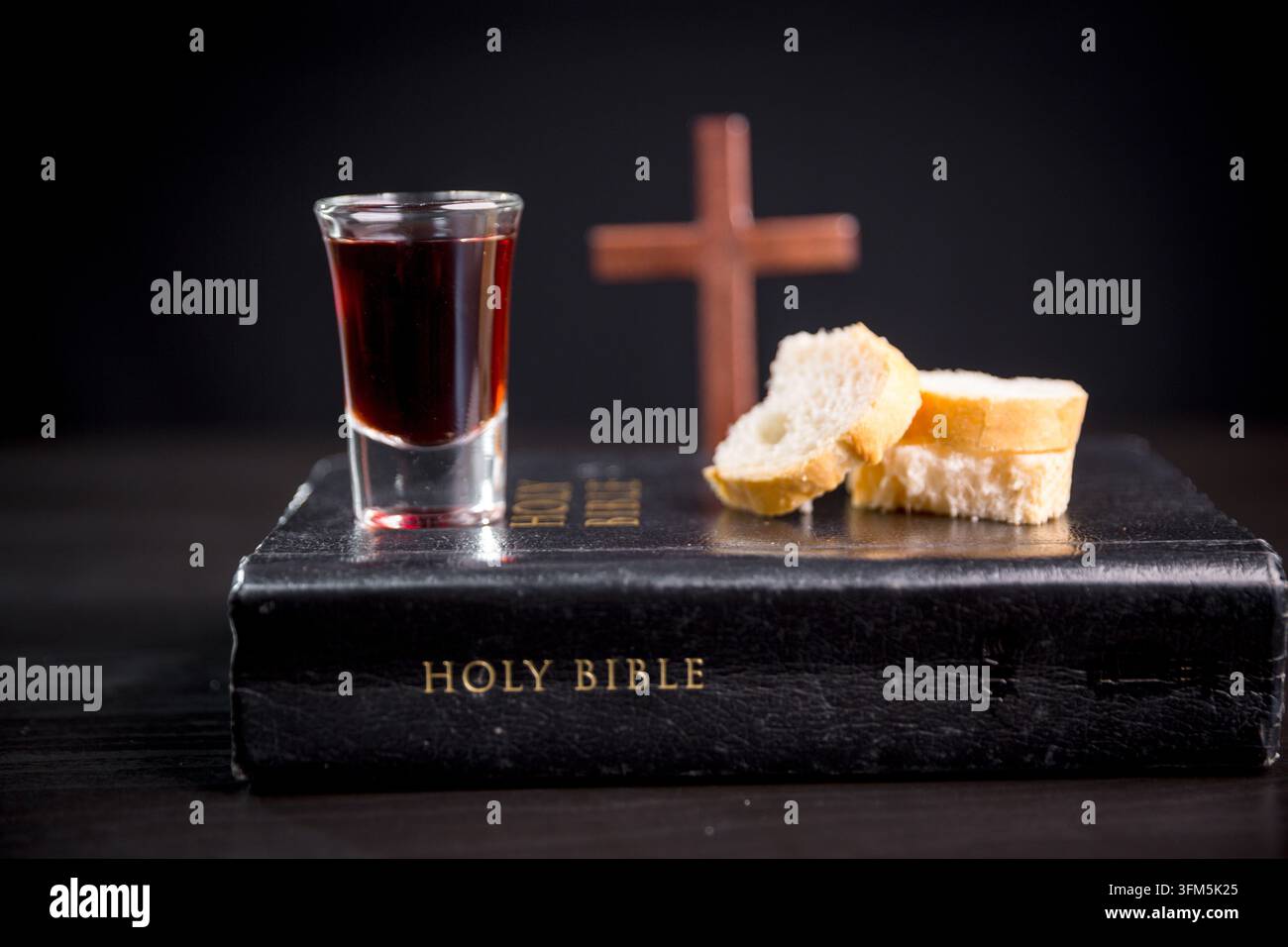 Dunkles rotes Getränk und geschnittenes Brot, arrangiert auf einem geschlossenen religiösen Buch mit einem hölzernen Kreuz im Hintergrund, das die christliche Kommunion, das Bibelstudium illustriert Stockfoto