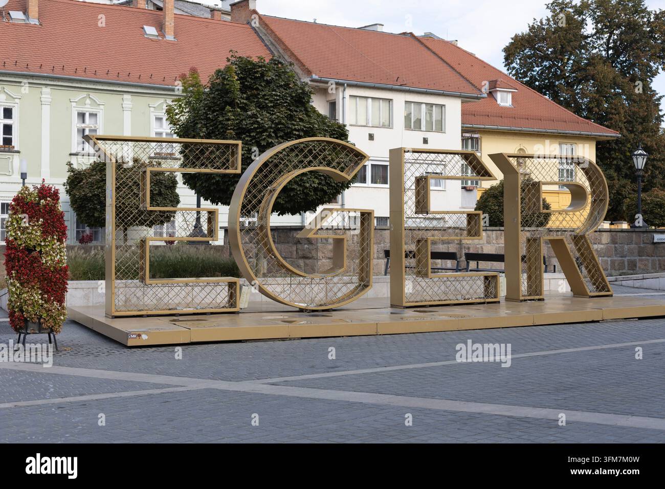 Große dekorative Eger-Schriftzüge stehen auf einem gepflasterten öffentlichen Platz, mit historischen Gebäuden, Bäumen, Blumenkästen und einer Straßenlaterne im Hintergrund. Tageslicht Stockfoto