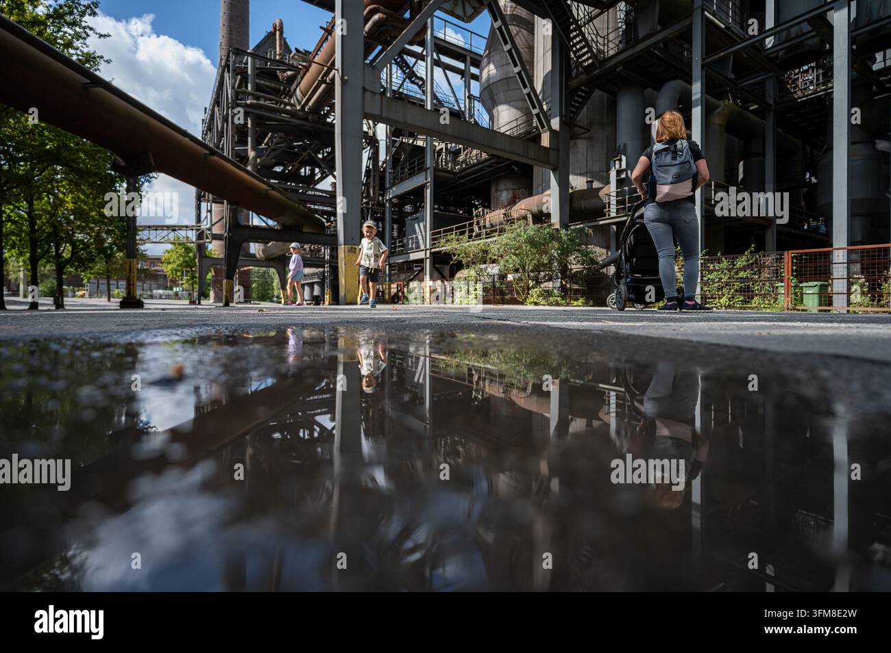 Kinder und ein Erwachsener gehen zwischen Stahlkonstruktionen, die sich in einer Pfütze im Landschaftspark Duisburg-Nord, Deutschland, spiegeln Stockfoto