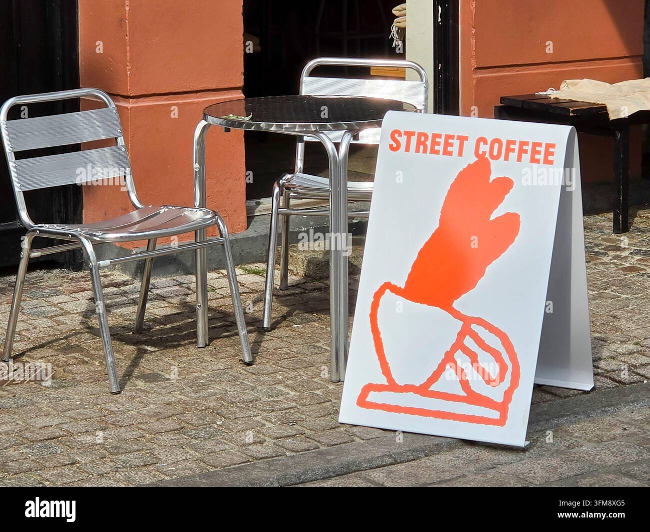 Weißes und orangefarbenes Gehweg-Schild / A-Frame-Schild mit dem Text „Straßenkaffee“ und einem Bild einer Kaffeetasse auf einer Straße; neben Metallstühlen und einem Tisch Stockfoto