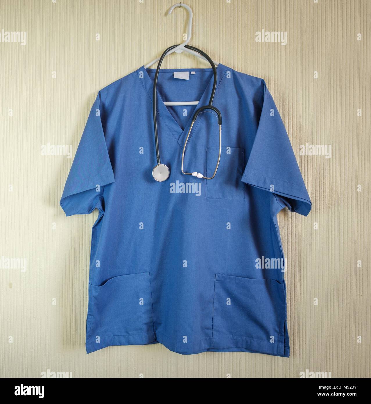 Blaues medizinisches OP-Kittelhemd und Stethoskop, die an einem Plastikhanger an einer strukturierten Wand hängen Stockfoto